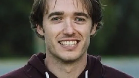 A smiling man with light brown hair wearing a maroon hoodie outdoors.