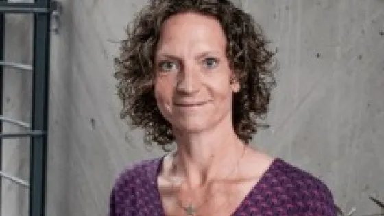 A woman with curly hair, wearing a purple patterned top, stands smiling against a gray concrete background.