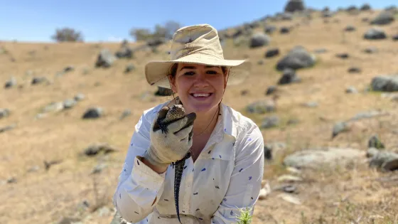 A smiling woman in a hat and gloves holds a lizard, sitting in a grassy field with large rocks scattered around.