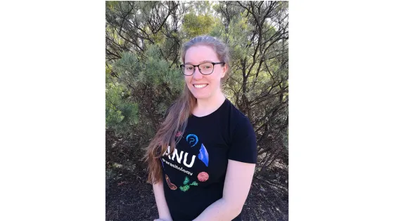 A woman with glasses and long hair, wearing a black t-shirt, smiles in front of a backdrop of green trees and bushes.