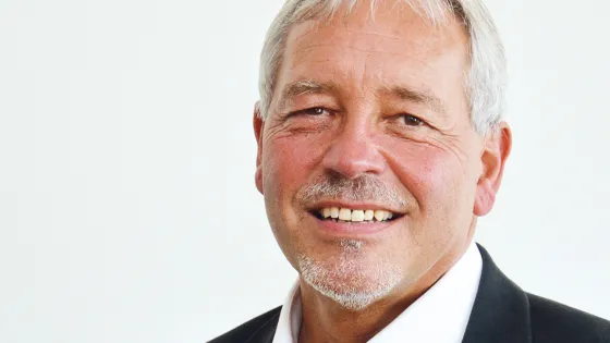 A portrait of a smiling older man with a white mustache and graying hair, wearing a black suit and white shirt.