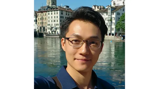 A young man smiles for a selfie with a scenic river and traditional European buildings in the background.