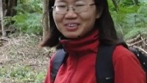 A woman in a red jacket and glasses smiles while standing in a forest.