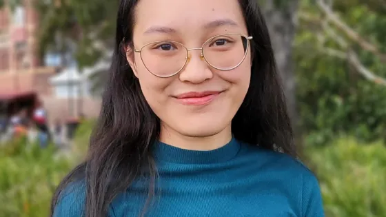 A young woman with glasses, wearing a teal top, smiling in front of a blurred natural background.