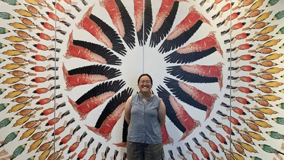 A woman smiling in front of a colorful wall display featuring a radiating pattern of feather illustrations.