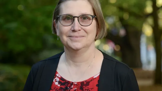 A woman with glasses wearing a black jacket and a red floral blouse smiles gently in a park setting.