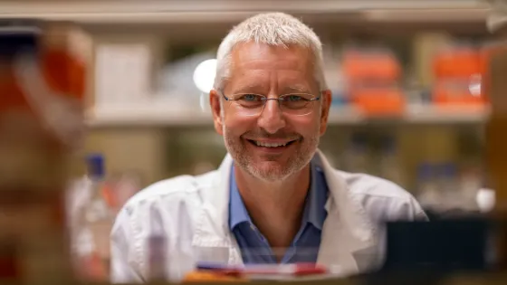 A smiling man with glasses wearing a white lab coat in a laboratory setting.