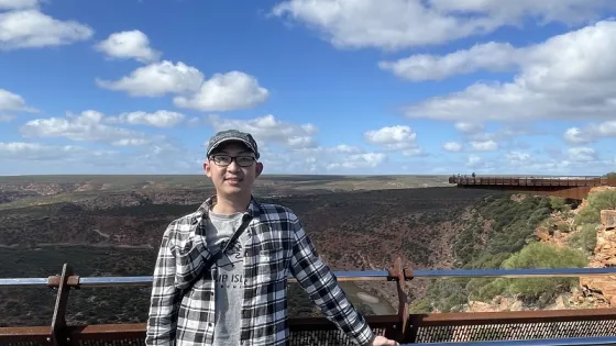 A person wearing a cap and glasses stands at a viewpoint, with a scenic view of a canyon and a cloudy sky in the background.