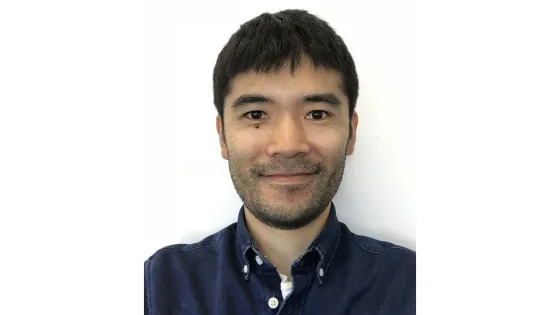 A headshot of a smiling man with short black hair, wearing a blue denim shirt, against a plain white background.
