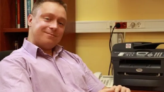 A man in a pink shirt smiling while sitting in an office chair next to a desk with a printer.