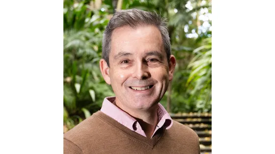 A smiling middle-aged man wearing a brown sweater over a pink shirt, standing in front of lush greenery.