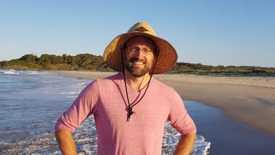 A man in a pink shirt and straw hat stands smiling on a sunny beach.