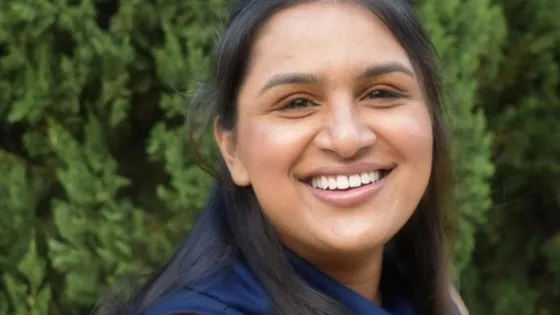 A woman with long dark hair smiling outdoors with green foliage in the background.