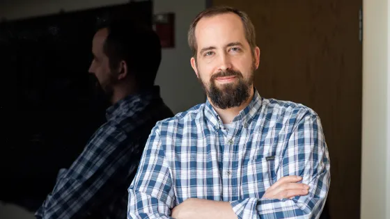 A man with a beard, wearing a blue plaid shirt, smiles slightly while standing with folded arms and his reflection visible in a dark glass window.