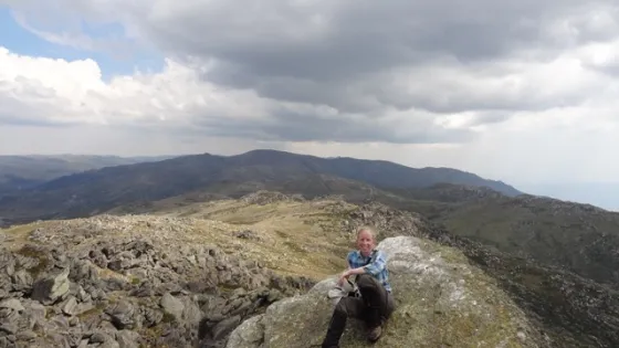 A person sits on a large rock in a mountainous landscape, surrounded by expansive, rocky terrain under a cloudy sky.
