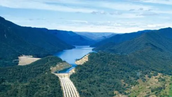 Aerial view of a dam with open spillways in a lush, mountainous landscape with a reservoir extending into the distance.
