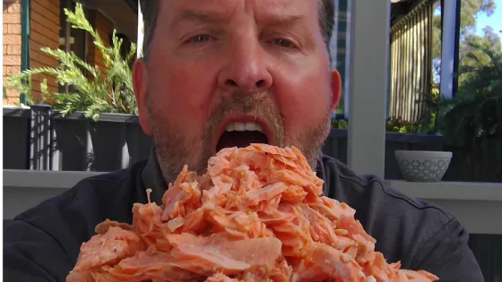 A man appears surprised by a large pile of salmon on a plate held close to his face, in an outdoor setting.