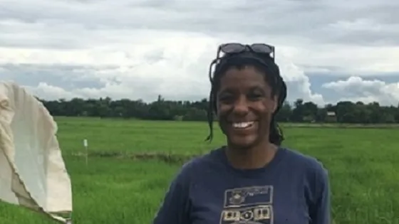 A woman smiling in a green field holding a butterfly net, with cloudy skies in the background.