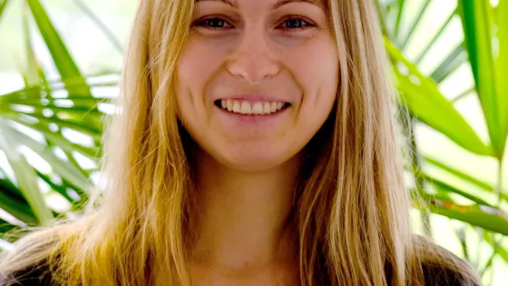 A smiling woman with long blonde hair, wearing a black shirt, standing in front of a green, leafy background.