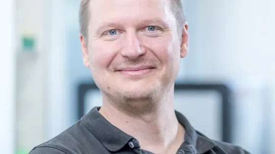 A man with light brown hair smiling at the camera in an office environment.