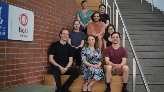 A group of eight diverse people smiling and sitting on steps at the Bio21 Institute.