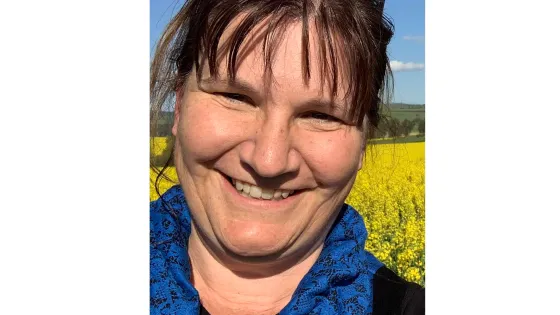 A smiling woman with sunglasses on her head standing in front of a vibrant yellow rapeseed field under a clear blue sky.