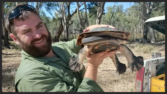 A smiling man in a green shirt holds a large turtle outdoors.