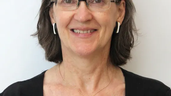 Portrait of a smiling woman with glasses, wearing a black shirt and small earrings, against a white background.
