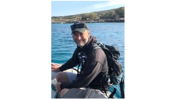 A man wearing a cap and backpack smiling while sitting on a boat with the sea and coastline in the background.