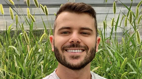A smiling man with a beard stands in front of tall green plants in a greenhouse.