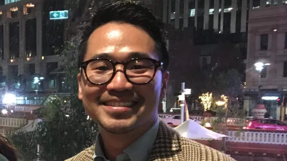 A smiling man wearing glasses and a patterned jacket stands in front of a cityscape at night.