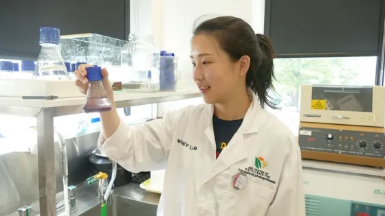 A scientist in a lab coat examines a flask of purple liquid in a laboratory setting.