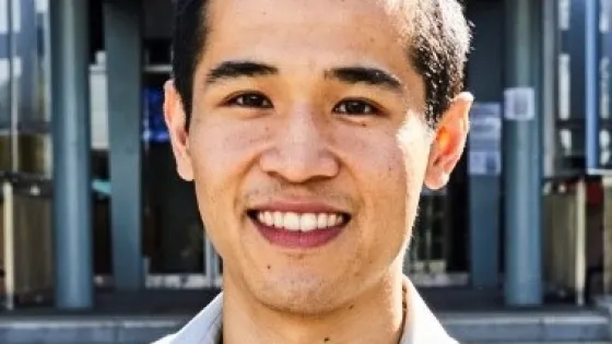 A smiling young man with short black hair in a gray shirt standing in front of a building.