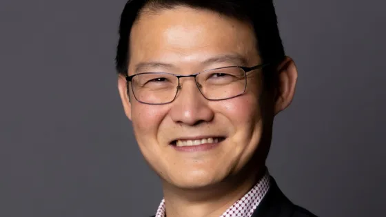 A professional portrait of a smiling Asian man wearing glasses, a checkered shirt, and a dark jacket.