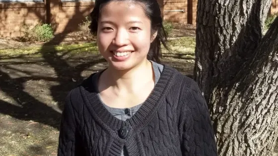 A young woman smiles brightly while standing outdoors in front of a tree and a brick building on a sunny day.