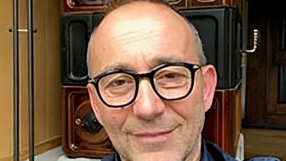 A man with glasses smiling at the camera, seated in front of a decorative wooden door.
