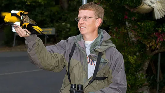 A person in a jacket with binoculars smiles while holding a colorful bird on one hand, as another bird flies nearby.
