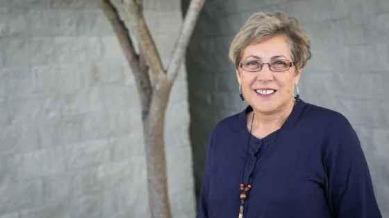 An older woman with short gray hair, wearing glasses, a dark blue blouse, and a beaded necklace, smiles while standing in front of a gray wall with a tree branch to the left.