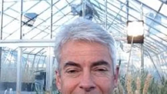 A man smiling in a greenhouse with tall plants in the background.