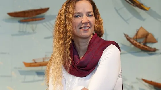 A woman with curly blonde hair smiles warmly while wearing a white blouse and red scarf, with a background featuring a map and model boats.