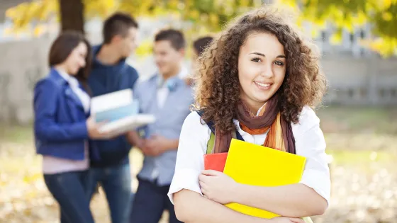 A smiling young woman with curly hair holds textbooks and stands outdoors, with three other students blurred in the background.