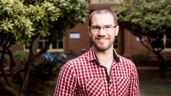 A man in glasses wearing a red plaid shirt smiles in front of a brick building and green shrubbery.
