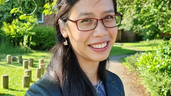 A smiling woman with glasses, wearing a black jacket, standing in a sunny park with green trees in the background.