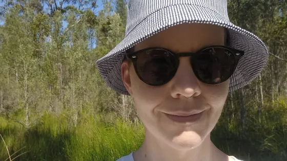 A person wearing a striped hat and sunglasses stands in front of a lush green forest.