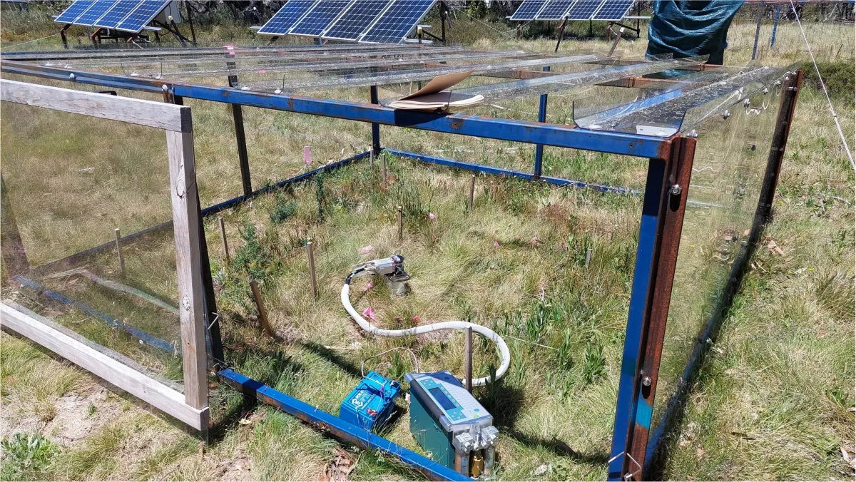 An outdoor experimental setup with solar panels and scientific equipment surrounded by a wire mesh fence in a grassy field.