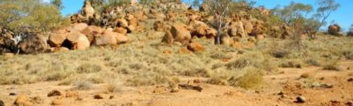 A dry, sparse landscape featuring red rocky outcrops and scattered scrub under a clear blue sky.