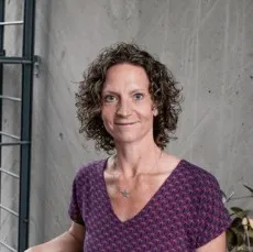 A woman with curly hair, wearing a purple patterned top, stands smiling against a gray concrete background.