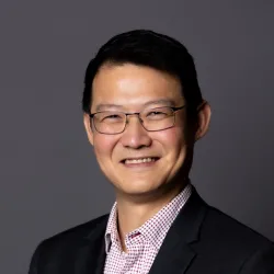 A professional portrait of a smiling Asian man wearing glasses, a checkered shirt, and a dark jacket.