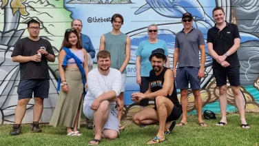 A group of nine diverse people smiling and posing in front of a colorful mural outdoors.
