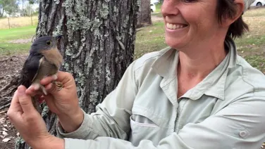 A smiling woman in a hat and outdoor shirt gently holds a small bird near a tree.